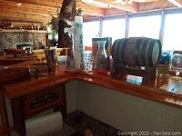 Bar counter setup with assorted barware showing yellow margarita glasses, plastic cups stacked high, glass decanter, and small wooden barrel on stand.