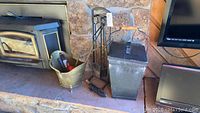 Fireplace tools, ash bucket, metal bucket with tools, and black firewood rack next to stone fireplace