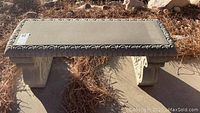 Concrete rectangular bench with ornate trim and carved leg supports in outdoor setting with brown pine needle ground cover