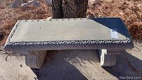 Concrete bench with ornate scroll design on sides and decorative edge pattern, placed outdoors on ground with natural light.