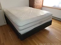 View of full bed with mattress on metal frame, showing mattress quilted white cover with gray sides, in room with hardwood floor and dresser in background.