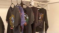 Photo showing side view of five t-shirts hanging on hangers. Mostly dark colored with different prints visible on front, confirming variety of designs.
