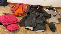 Photo of colorful ski outerwear including orange jacket and pants, pink pants, black jacket, and sweaters laid out on carpet