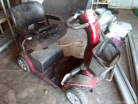 Red Pride electric mobility scooter in a garage setting showing the full left side including the seat, steering column, and front basket. Dust and minor wear visible.