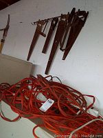 Orange extension cords and white surge protectors piled on a surface with tree pruner pole and hand saws visible on wall.