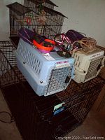Photo showing large blue Pet Taxi plastic pet crate with wire door, white pet carrier, and brown metal bird cage with assorted pet collars, muzzles, and training collars on top