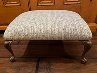 Front close-up view of the upholstered footstool showing textured woven fabric in neutral tones with brass cabriole legs
