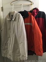 Three men's winter jackets hanging on hangers: beige, orange/black, red/black