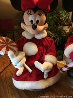 Close-up of Minnie Mouse Christmas figurine dressed in red velvet with white fur trim, holding a candy cane, showing details of clothing and expression.