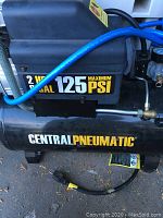 Side view of air compressor motor casing showing '2 HP' and '125 MAXIMUM PSI' labels and 'Central Pneumatic' branding on tank, power cord on ground.