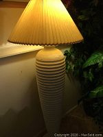 Tall beige lamp illuminated, showing pleated beige shade and plaster ring column base