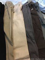 Photo showing 7 pairs of men's pants in various neutral colors including beige, tan, grey, brown, and dark shades.