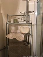 Photo showing silver metal bathroom storage rack with various accessories including a hand towel ring, magazine holder, in-tub bath mat, soaking pillow, ceramic water glass and soap dish, toothbrush holder, and a floor mat.