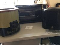 Photo showing the green crockpot, black toaster oven and black deep fryer on a white table surface.
