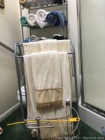 Over the toilet shelving with towels stacked and rolled on the shelves, free standing towel rack with hanging towels, yellow hair removal tool placed at base.