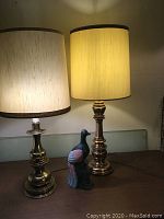 Full view showing both brass lamps with beige fabric shades and hand carved wooden bird figurine on table.