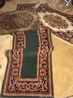 Three different rugs placed on the floor: a green and black outlined runner with rust floral patterns, a beige and brown oval rug with floral design, and a beige, green and rust area rug.