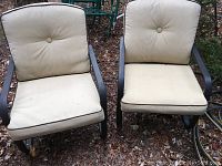 Front view of two outdoor metal chairs with beige cushions and button tufting, placed on ground with leaves.
