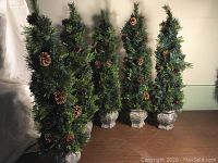 Group photo of five artificial evergreen trees in rough cement-style pots, decorated with pinecones and some lights not working.