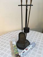 Set of three metal fireplace tools placed in metal holder, showing shovel, poker, and brush with black bristles. Two unopened boxes of long matches in different decorative boxes seen to the side.