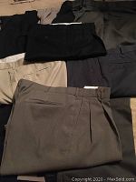 Photo showing nine pairs of men's dress pants arranged flat, displaying various colors and typical dress pant details such as side and back pockets.