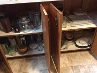 Open kitchen cabinet shelves holding various glass bakeware dishes including Pyrex clear glass and ceramic pieces, some with lids, assorted bowls, pots, and kitchen items.