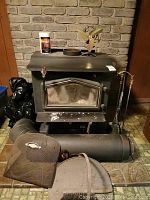 Full view of black cast iron wood burning stove with attached stove pipe, stove fan on top, tools and mesh guard in front.