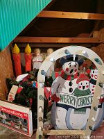 Wooden snowmen holding 'Merry Christmas' sign with mounted lights, inflatable lit candles, and some greenery visible behind