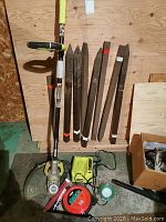 Ryobi weed trimmer with battery charger, red and green spools of twine, and six wooden garden stakes leaning against plywood background on concrete floor.