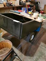 Side view of black steel utility dump trailer cart with many household items on the bed, showing pneumatic tires and towing arm.