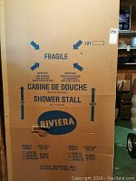 Photo of large cardboard box packaging for Riviera shower stall, model DPB-32 white, with fragile markings and product information.