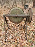 Photo shows front view of the old iron cement mixer with large gear and stand on leafy ground