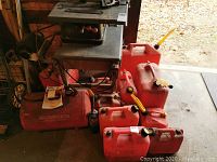 Group of red plastic gas cans in assorted sizes, some fitted with yellow spouts, arranged on a workshop floor near tool equipment.