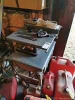 Front and top view of the vintage heavy-duty table saw with tools and extension cord on top and red fuel containers underneath.