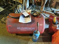 Red Motomaster portable air tank with visible brand and capacity markings. Air tank resting on floor with some dirt and rust.