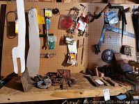 Overall view of wall-mounted pegboard holding various hand tools and hardware, with a wooden bench surface below covered with more tools including clamps, scrapers, and wrenches.