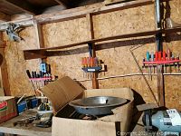 Wide view showing cardboard box containing various tools and hardware, shelves with screwdrivers with colored handles, round metal tray, and other miscellaneous items.