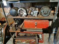 Four electric motors on a top shelf showing rust and wear, older gear pumps, red closed toolbox, and an open toolbox with assorted hand tools.