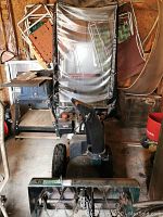 Front view of green and black two-stage snowblower with rusted auger housing, black adjustable chute and operator canopy in shed.