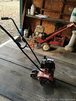 Full view of the red Murray rototiller with handles, wheels, and tines on concrete floor inside a shed, showing the unit and surrounding environment.