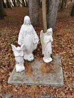 Photo shows three white stone religious statues shaped as Virgin Mary and angels set on a rectangular concrete slab surrounded by brown fallen leaves and trees in background.