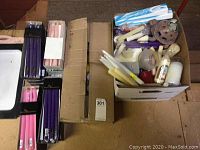 Photo shows multiple boxes, including unopened boxes of taper candles in purple, pink, and other colors alongside a box with loose candles and candle holders.