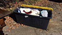 Black plastic storage bin with yellow lid containing folded men's clothing including dress shirts, sweaters, and tie.