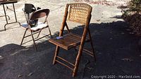 Front view of light brown rattan folding chair outdoors on pavement, showing woven cane backrest and slatted seat
