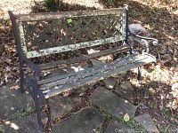 Full view of wrought iron bench showing overall condition, with wood slats largely deteriorated and bench placed outdoors on stone slabs with surrounding leaves.