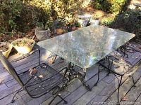 Four black cast iron outdoor chairs arranged around a glass top table on a wooden deck, showing two sizes of chairs with mesh seats and leaf decoration.