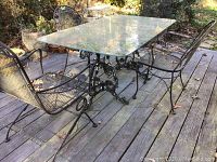 Rectangular cast iron table base and glass top shown on wooden deck outdoor setting, with matching iron chairs in background not included in lot