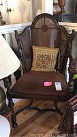 Full view of wooden chair showing carved details on arms and legs, woven cane back and sides, brown upholstered seat cushion, floral pillow on seat, sitting on hardwood floor.