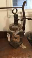 Full view of vintage glass butter churn with metal lid, hand crank and wooden paddle inside jar, placed on wooden surface.