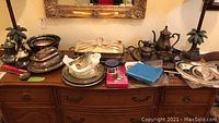 Various silver plated serving items displayed on a wooden sideboard, showing a range of platters, bowls, and lidded dishes with a mirror in the background